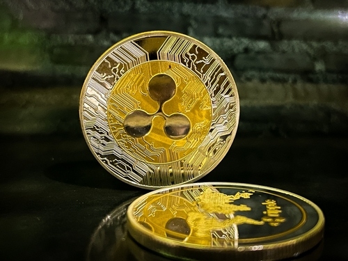A close-up photograph of two XRP (Ripple) cryptocurrency coins, one standing upright showing the logo and another lying flat, resting on a dark surface.