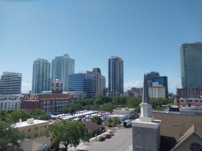 A sunny aerial view of downtown St. Petersburg, Florida, showcasing its skyline with modern high-rises, a church steeple in the foreground, and what appears to be an outdoor festival or event with tents set up along the street, setting the scene for a fun birthday weekend celebration in the city.