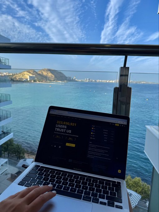 A person uses a MacBook Air displaying a Binance login page with the text '323,930,921 USERS TRUST US', set against a scenic coastal view.
