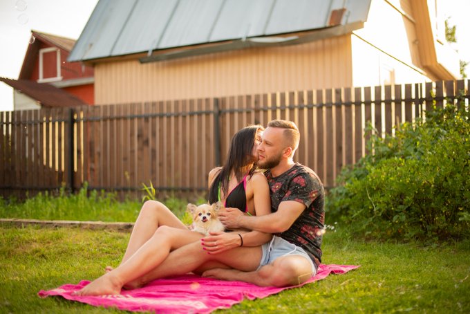 A couple sitting on a pink blanket in a grassy yard holding a small white dog, with a wooden barn and fence in the background.