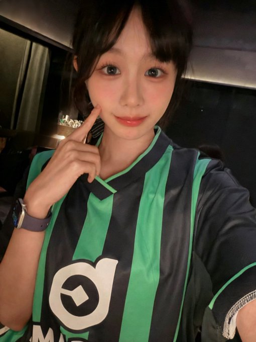 The image features a young woman wearing a green and black striped shirt, posing for a photo with her finger on her nose. The background of the picture shows a bus interior, which adds an interesting context to the scene. The caption accompanying the image reads "想不想跟俺穿情侣装 两层欠球衣 @bcgame". This suggests that the woman might be attending or participating in a bus-themed event, possibly related to sports.