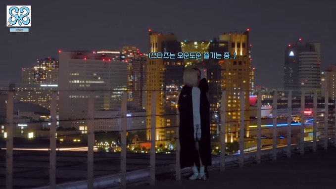 A person in a long black coat stands on a rooftop at night overlooking a brightly lit city skyline, with Korean subtitles reading '(스타는 오른모로 즐기는 중..)' and a 'GO ON MEDIA' logo in the top left corner.
