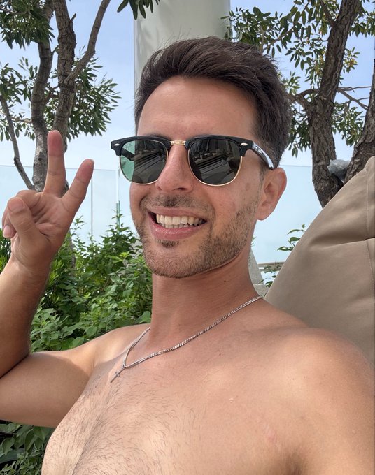 A shirtless man wearing sunglasses smiles and holds up a peace sign in an outdoor setting with green foliage and a bright sky in the background.