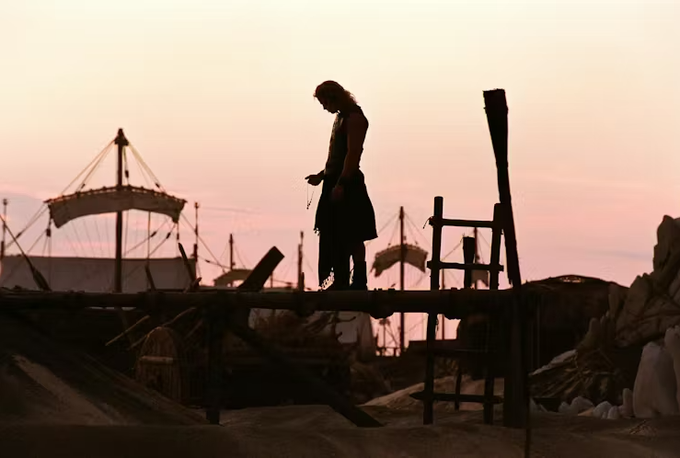 A silhouetted figure in a kilt stands on a wooden pier at sunset holding a chain, with large ship structures visible in the background.