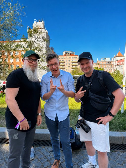 A photograph of three men posing outdoors in an urban setting with a tall building in the background; the man in the center is making rock-on hand gestures.