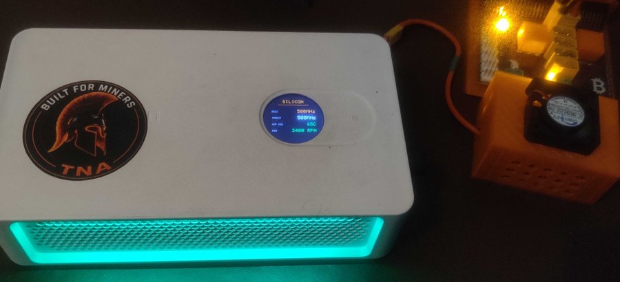A cryptocurrency mining device with 'BUILT FOR MINERS TNA' branding displays real-time hashrate statistics (500kHs) on its small screen.