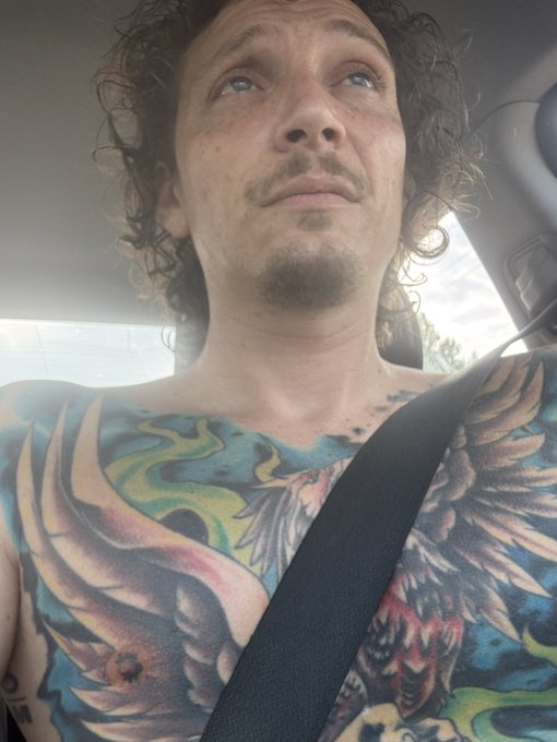 A shirtless man with curly hair, a goatee, and extensive chest and arm tattoos sitting in a car with a seatbelt on.
