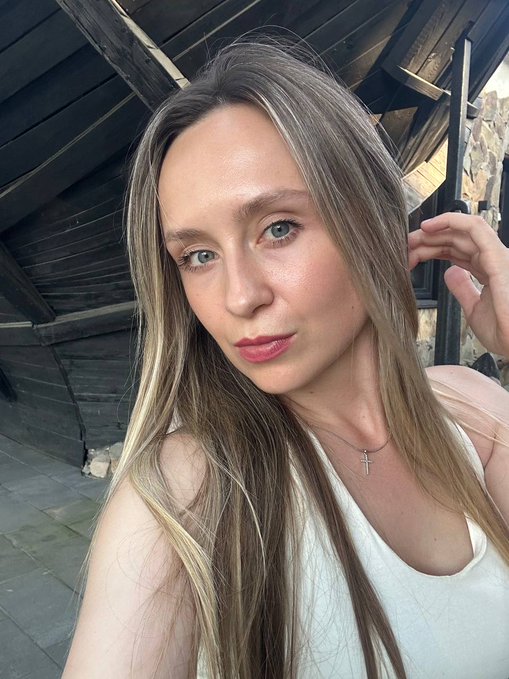 A selfie of a young woman with long blonde hair, wearing a white top and a cross necklace, with a wooden slatted ceiling in the background.
