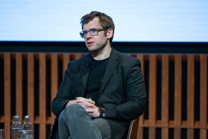 A man with glasses and a beard, wearing a dark blazer over a black shirt, sits on stage with hands clasped during a panel discussion at the Goldman Sachs Digital Asset conference.