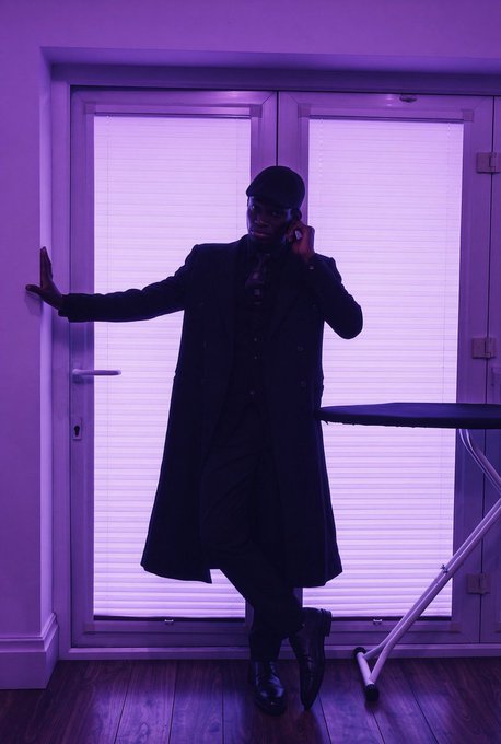 Stylized photo of a man in a long dark coat and cap leaning against a door with purple lighting and an ironing board beside him.
