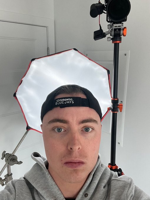 A selfie photo of a young man wearing a backwards 'TORONTO BLUE JAYS' cap and grey hoodie, with professional photography lighting equipment (octabox softbox and light stand) visible behind him.