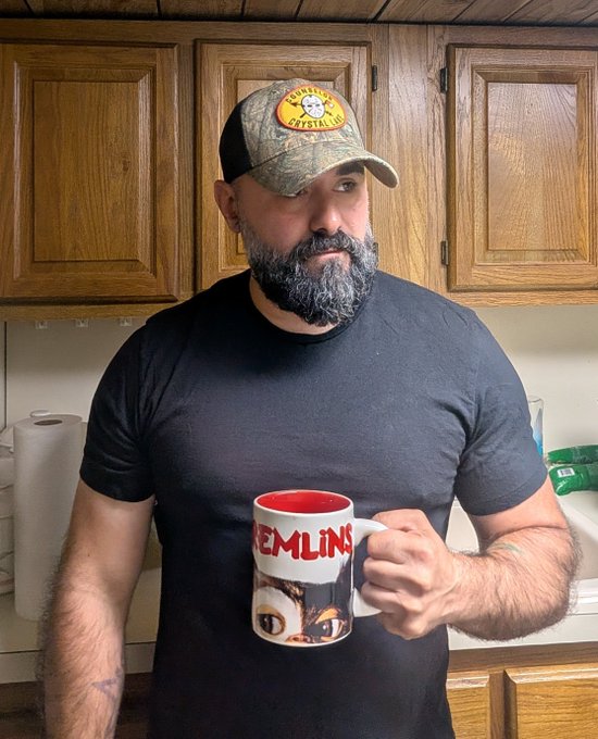 A bearded man in a black t-shirt and baseball cap holds a mug featuring 'GREMLINS' text and Gizmo's face, with his own eyes edited to match the character.