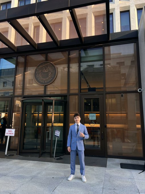 A young man poses in front of the Federal Reserve building in Washington, D.C., wearing a blue suit and displaying the new Morpho Merch, with a "100% ID CHECK" sign visible near the entrance.