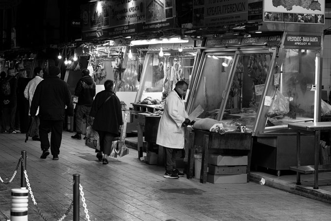 A black and white photograph of a bustling indoor market with butchers and vendors at counters displaying meat under glass cases, with signs visible including 'CRETA ΠΡΕΒΕΖΑ' and 'ΒΕΛΛΑΣ ΚΑΛΛΗ ΣΦΑΓΕΙΟ'.