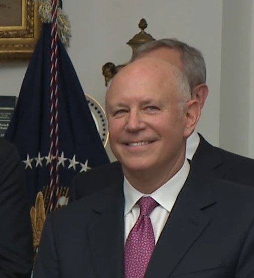 A smiling older man in a dark suit and pink patterned tie, photographed in an official setting with an American flag and presidential seal visible in the background.