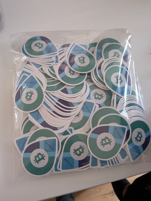 The image shows a collection of small, square stickers with blue and green colors, each featuring a white Bitcoin symbol. These stickers appear to be part of a promotional campaign or product packaging for Bitcoin. The stickers are neatly arranged in rows on a table, showcasing their design and purpose.