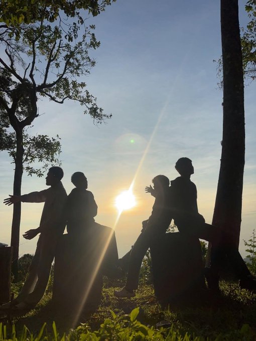 Four silhouetted figures pose dramatically against a sunset sky framed by trees.