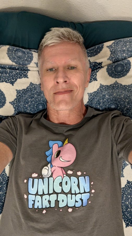 A selfie of a gray-haired man lying on a bed with blue-patterned bedding, wearing a dark gray t-shirt featuring a pink unicorn and the text 'UNICORN FART DUST'.
