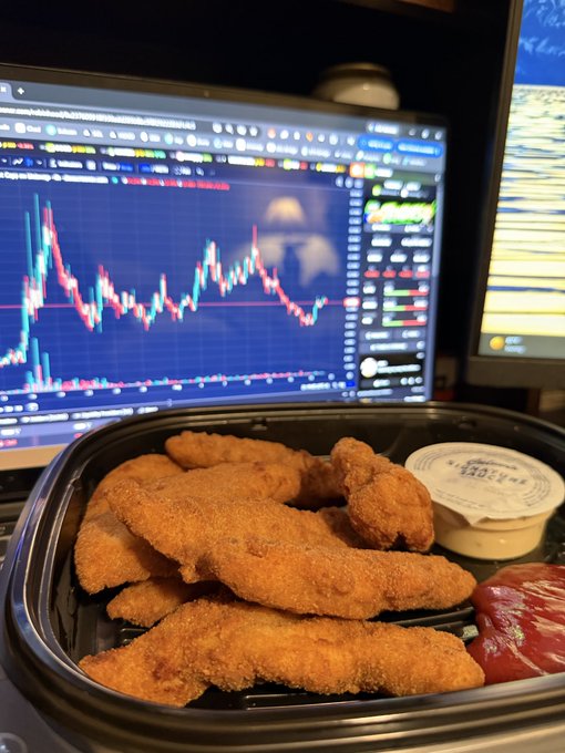 A container of chicken tenders sits in front of a laptop displaying a cryptocurrency price graph on a trading platform.