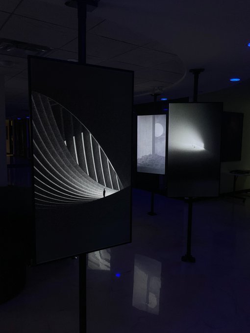 A dimly lit modern art exhibition space featuring illuminated display panels showing black and white photographs, including an image of a curved architectural structure with a small figure.