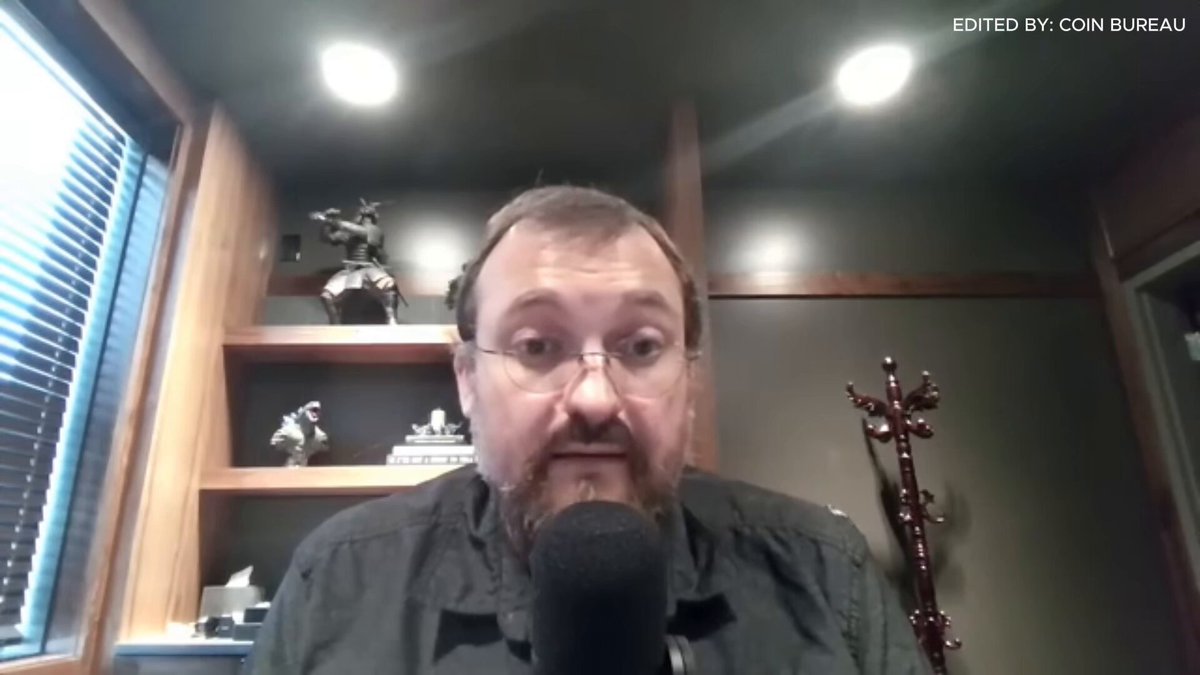 The image shows a man wearing glasses sitting behind a microphone, possibly recording a podcast or live stream. The background features a green wall with a window that allows natural light into the room. There are also several books scattered around the space, suggesting an intellectual atmosphere. A red coat is hanging on a hook in front of the man, adding a pop of color to the scene.