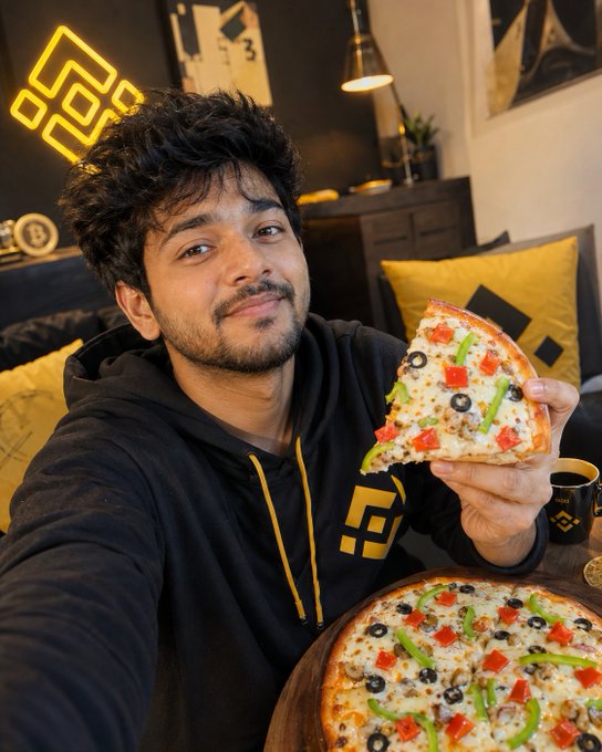 In the image, there's a man holding up half of a pizza with his left hand, as if he's taking a photo or making a video. The pizza has various toppings including pepperoni, olives, cheese, and peppers. There is also a coffee cup near him that matches the color scheme of the image. The image has a Binance hashtag on it which indicates a possible connection to cryptocurrency, and there are charts displayed in the background suggesting that this could be a promotional post or tutorial related to the Binance platform.