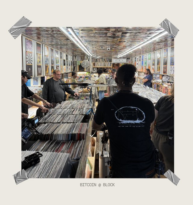A bustling record shop filled with vinyl crates and posters covering every wall, where several patrons browse the bins—including a man in a black "Bitcoin @ Block" t-shirt capturing the scene with his camera, fittingly illustrating the "In the lab" theme of hands-on discovery and curation.