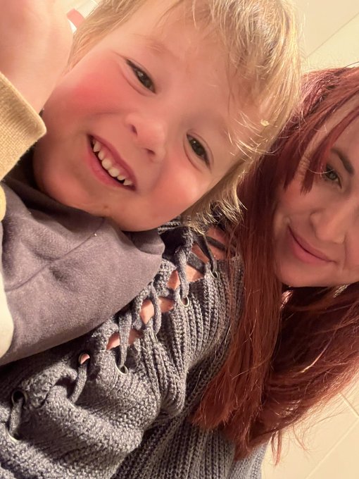 A close-up selfie of a smiling blonde child and a woman with long red hair, both wearing grey knit sweaters.