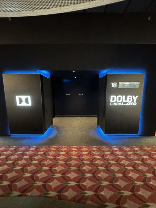 An entrance to a theater featuring a Dolby Cinema at AMC auditorium, marked by the illuminated "DO" logo on the left and "18 DOLBY CINEMA at AMC" signage on the right, set against a patterned red carpet.
