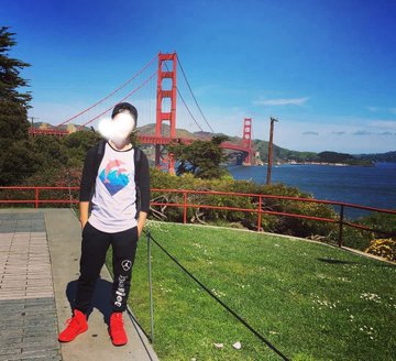 A young person wearing a white t-shirt with a blue heart design stands on a grassy area in front of the Golden Gate Bridge on a sunny day.