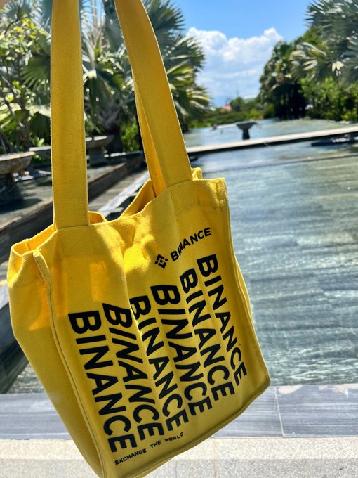A bright yellow tote bag covered in repeating 'BINANCE' text with the tagline 'EXCHANGE THE WORLD,' photographed outdoors near a pool with tropical plants.