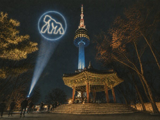 The image shows a night scene at N Seoul Tower in South Korea, where a spotlight projects the Bored Ape Yacht Club (BAYC) logo—an illuminated ape silhouette—into the sky above the illuminated pagoda and tower. This visual spectacle ties into the post about "Seoul is waking up" with $APE, likely referencing an APE token promotion or event connected to the Bored Ape NFT collection.

No specific text, numbers, or tickers appear beyond the BAYC logo symbol itself.
