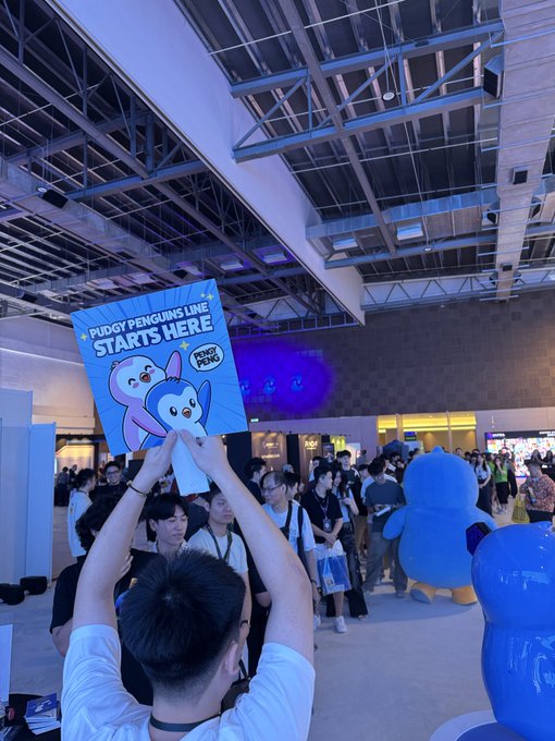 A person holds up a sign reading "PUDGY PENGUINS LINE STARTS HERE" above a crowd of attendees gathered at what appears to be a Pudgy Penguins event, directly illustrating the lengthy queue mentioned in the caption.