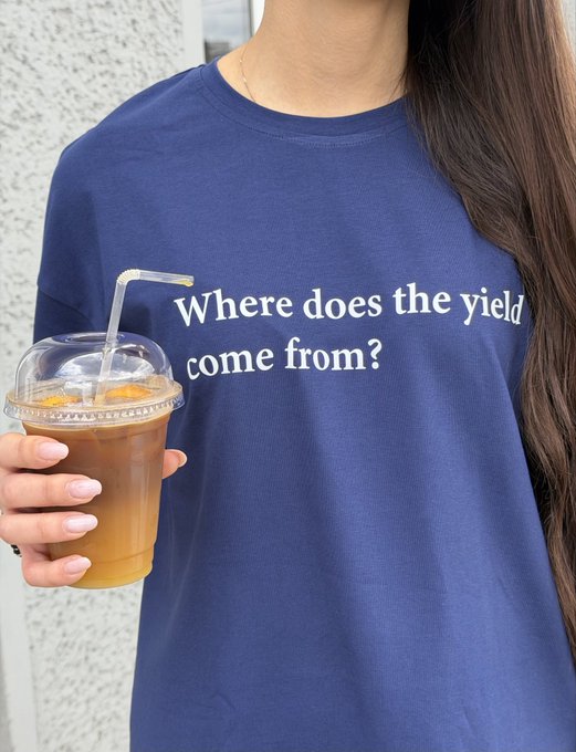 A person wearing a navy blue t-shirt with the text "Where does the yield come from?" while holding an iced coffee drink in a clear plastic cup.