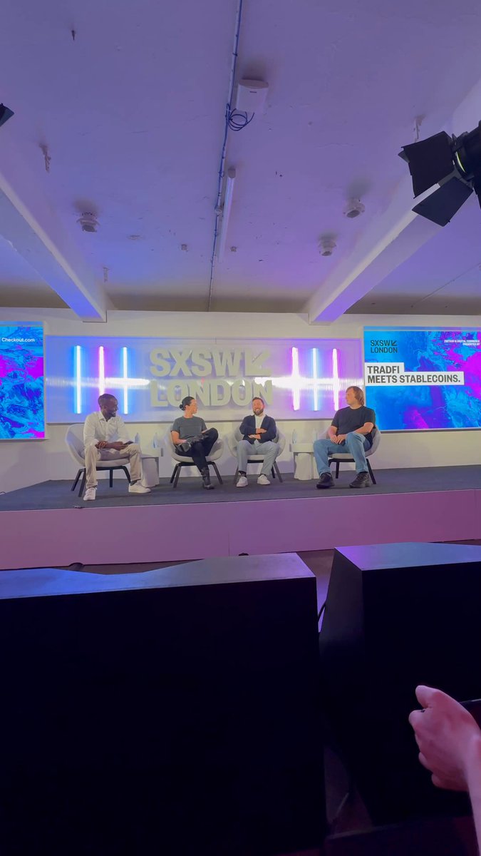 The image shows a group of people sitting around a table, with one person holding up their phone screen displaying a message from @kmgrose UK CEO of @coinbase, discussing stablecoins as a growing trend in payments. The scene takes place at an event or gathering where the participants are engaged in conversation and sharing ideas about this emerging concept.