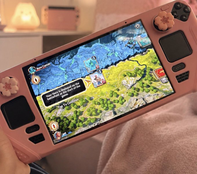 A hand holds a pink handheld gaming console displaying "Born Captain" on its screen, with a tutorial prompt reading "Your Hero is Deployed on the battlefield. It is the start of the game. Tap To Continue," set against a cozy background with a small lamp and game console. The image supports the post "Proof of gaming — Only on @Immutable" by showcasing a vibrant mobile gaming experience on a cute, custom-colored device.