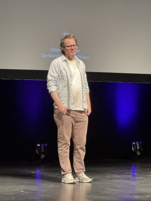 Charlie, co-founder of Expo (visible in the partial text on the screen behind him), stands on stage in a casual outfit of an unbuttoned light shirt, beige pants, and sneakers, kicking off ChainReactConf as referenced in the post.