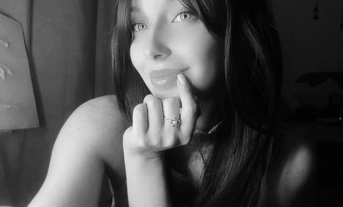 Black and white photo of a woman with long dark hair posing with her finger on her lip, showing a ring on her finger in a softly lit room.
