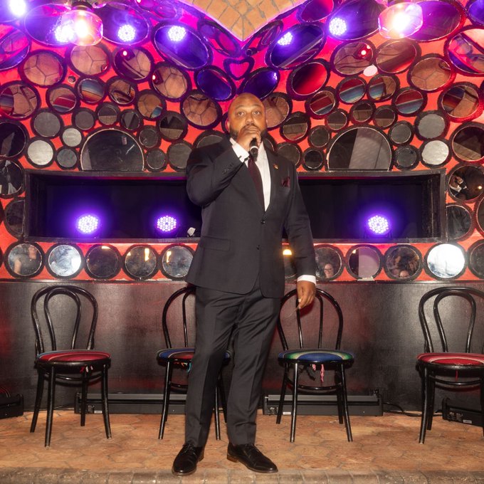 A man in a dark suit with a red tie stands on a stage holding a microphone, backed by a large metallic wall decoration with circular mirrors and purple stage lighting.