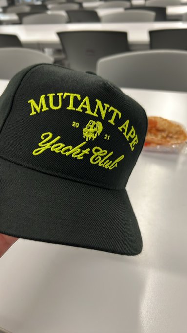 A dark green baseball cap with neon yellow embroidery reading "MUTANT APE YACHT CLUB 2021" rests on a table in what appears to be a classroom or meeting room.