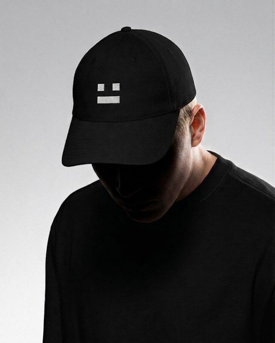 A minimalist portrait of a person wearing a black baseball cap with a simple, blocky face logo, and a black t-shirt, set against a gray background.