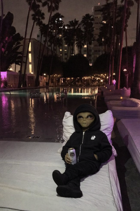 A child dressed in an alien costume and black hoodie lounges poolside on a cushioned chaise lounge at night, holding what appears to be a beverage can, with palm trees and string lights glowing in the background—capturing a quirky "The Club >" vibe.