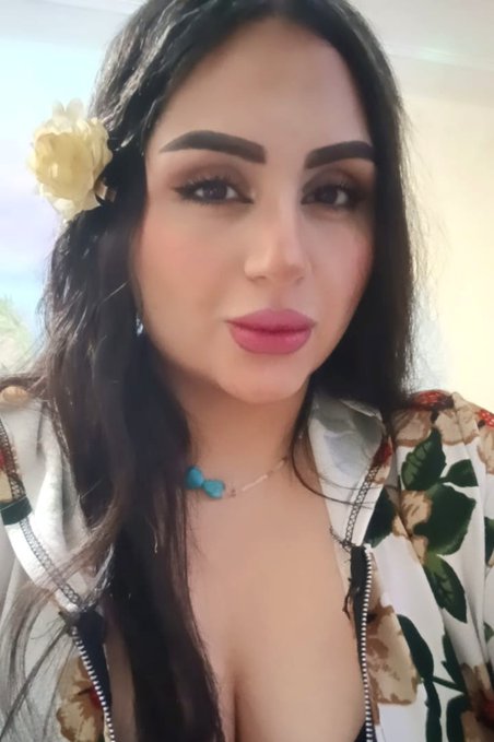 A close-up selfie of a woman with long dark hair, wearing a yellow flower hair accessory and a floral jacket, with a turquoise beaded necklace.