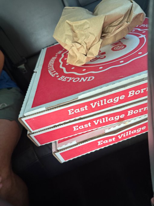 Three stacked "East Village Born" pizza boxes sit on a car seat, ready to be delivered to local "heroes" as a gesture of appreciation. A crumpled brown paper bag rests on top of the boxes.
