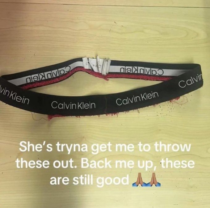 Photo of a worn black and red Calvin Klein waistband/underwear on a wooden surface, with text reading 'She's tryna get me to throw these out. Back me up, these are still good' with a prayer hands emoji.