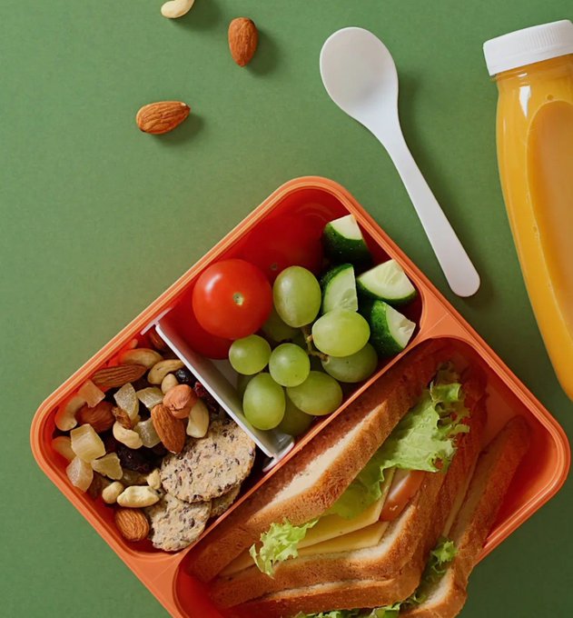 An orange bento lunch box containing a sandwich, cherry tomatoes, cucumber, green grapes, and mixed nuts, accompanied by a white spoon and an orange beverage bottle on a green surface.