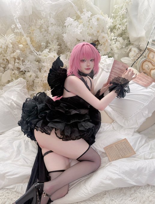 A cosplayer with pink hair and black wings poses on a white bed surrounded by white flowers, wearing a black ruffled dress and holding a sheet of music.