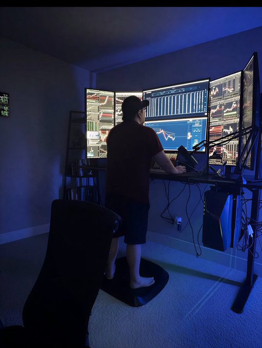 A trader stands barefoot on a mat at a multi-monitor trading setup displaying various charts and financial data in a dimly lit room with blue ambient lighting.