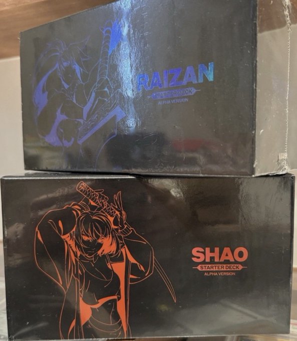Two sealed trading card game booster boxes stacked together: 'RAIZAN STARTER DECK ALPHA VERSION' in blue on top and 'SHAO STARTER DECK ALPHA VERSION' in orange/red on the bottom.
