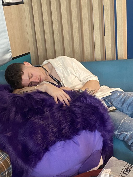 A person peacefully asleep on a teal couch, wearing a white shirt and lanyard, covered with a fluffy purple blanket.
